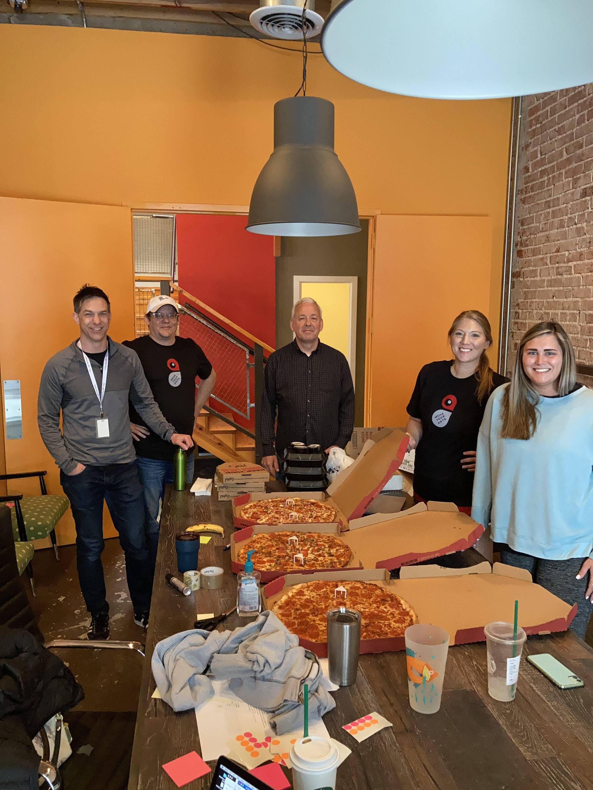 Group of people smiling around a table of pizza boxes.