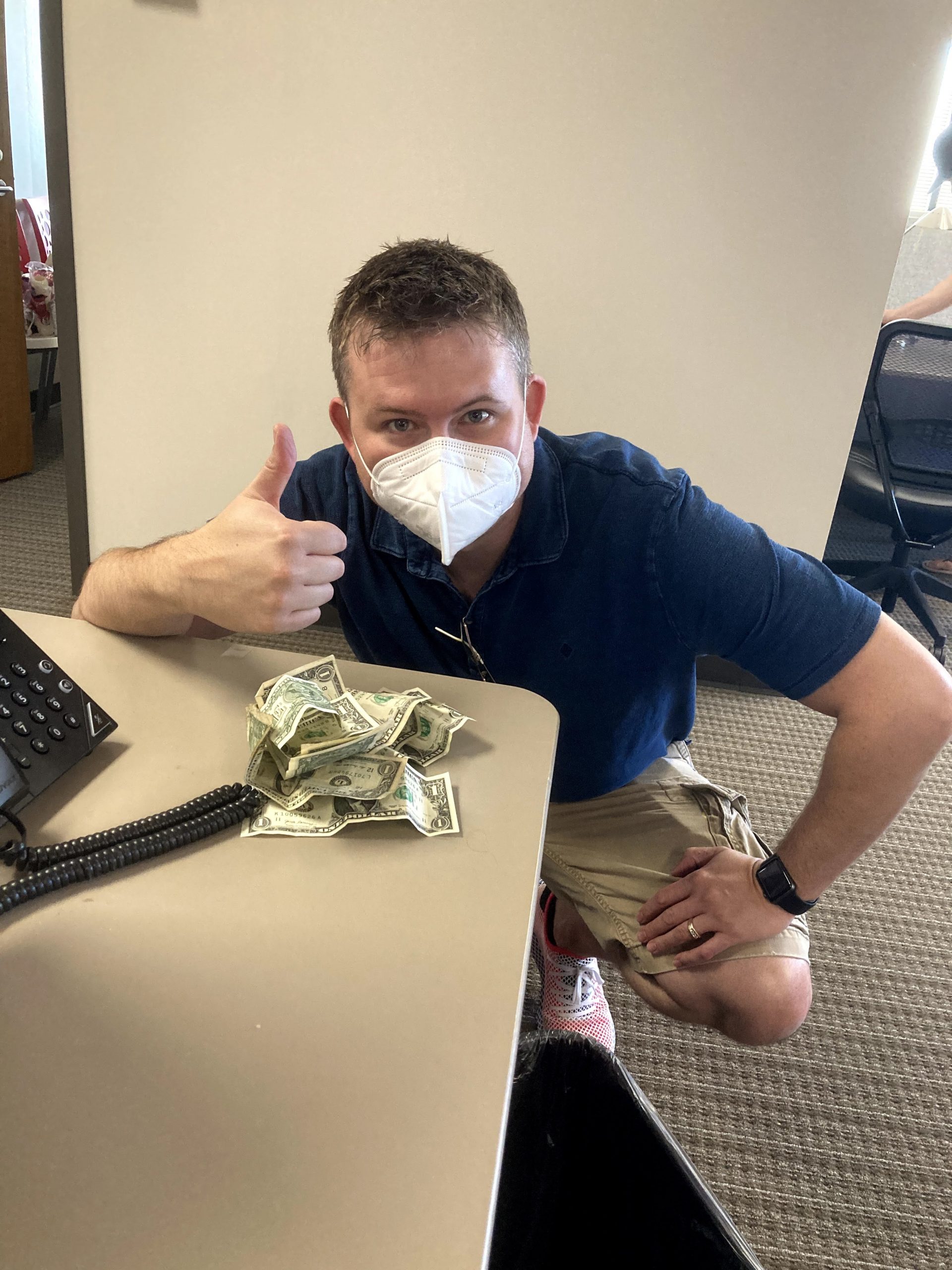 Man in mask making a thumbs up sign with hand next to a pile of dollar bills on a desk.